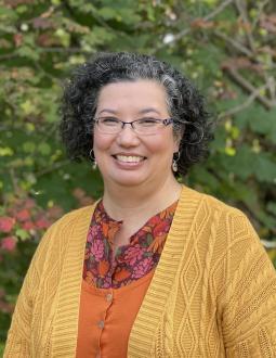 Head and shoulders shot of Sunni Nishimura. She smiles at the camera. She has chin length curly black hair and glasses. She is wearing fall colours (oranges, reds, and golds) and stands in front of a green tree.
